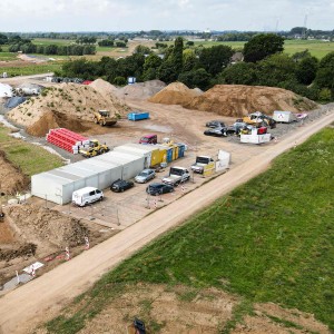 
Auf der linken Seite des Bildes ist eine Baustelle zu sehen, die von oben fotografiert wurde. In einem ausgehobenen Graben, der sich von links unten nach rechts oben erstreckt, sind mehrere Bagger und Bauarbeiter zu erkennen. Links im Vordergrund fährt ein großer Muldenkipper mit roter Kabine, beladen mit Erde, weg.
Im mittleren Bereich der Baustelle befinden sich mehrere große Erdhaufen und Baumaschinen. Daneben stehen eine Reihe von weißen und blauen Containern, die als Büros oder Unterkünfte dienen könnten. Davor parken mehrere Autos und Kleintransporter.
Der Hintergrund des Bildes zeigt eine ländliche Umgebung mit grünen Feldern und Bäumen. Ganz hinten ist eine Straße oder ein Damm zu erkennen. Der Himmel ist bewölkt.