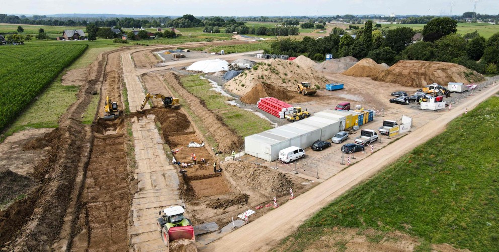 
Auf der linken Seite des Bildes ist eine Baustelle zu sehen, die von oben fotografiert wurde. In einem ausgehobenen Graben, der sich von links unten nach rechts oben erstreckt, sind mehrere Bagger und Bauarbeiter zu erkennen. Links im Vordergrund fährt ein großer Muldenkipper mit roter Kabine, beladen mit Erde, weg.
Im mittleren Bereich der Baustelle befinden sich mehrere große Erdhaufen und Baumaschinen. Daneben stehen eine Reihe von weißen und blauen Containern, die als Büros oder Unterkünfte dienen könnten. Davor parken mehrere Autos und Kleintransporter.
Der Hintergrund des Bildes zeigt eine ländliche Umgebung mit grünen Feldern und Bäumen. Ganz hinten ist eine Straße oder ein Damm zu erkennen. Der Himmel ist bewölkt.