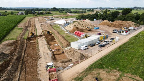 Auf der linken Seite des Bildes ist eine Baustelle zu sehen, die von oben fotografiert wurde. In einem ausgehobenen Graben, der sich von links unten nach rechts oben erstreckt, sind mehrere Bagger und Bauarbeiter zu erkennen. Links im Vordergrund fährt ein großer Muldenkipper mit roter Kabine, beladen mit Erde, weg.
Im mittleren Bereich der Baustelle befinden sich mehrere große Erdhaufen und Baumaschinen. Daneben stehen eine Reihe von weißen und blauen Containern, die als Büros oder Unterkünfte dienen könnten. Davor parken mehrere Autos und Kleintransporter.
Der Hintergrund des Bildes zeigt eine ländliche Umgebung mit grünen Feldern und Bäumen. Ganz hinten ist eine Straße oder ein Damm zu erkennen. Der Himmel ist bewölkt.