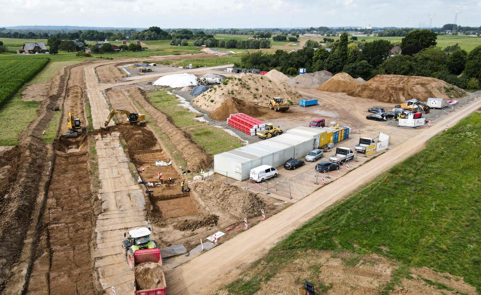 Auf der linken Seite des Bildes ist eine Baustelle zu sehen, die von oben fotografiert wurde. In einem ausgehobenen Graben, der sich von links unten nach rechts oben erstreckt, sind mehrere Bagger und Bauarbeiter zu erkennen. Links im Vordergrund fährt ein großer Muldenkipper mit roter Kabine, beladen mit Erde, weg.
Im mittleren Bereich der Baustelle befinden sich mehrere große Erdhaufen und Baumaschinen. Daneben stehen eine Reihe von weißen und blauen Containern, die als Büros oder Unterkünfte dienen könnten. Davor parken mehrere Autos und Kleintransporter.
Der Hintergrund des Bildes zeigt eine ländliche Umgebung mit grünen Feldern und Bäumen. Ganz hinten ist eine Straße oder ein Damm zu erkennen. Der Himmel ist bewölkt.