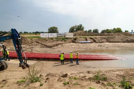 Ein Kran steht auf der linken Seite des Bildes. Ein langer, roter Rohrstrang, der in einer horizontalen Reihe auf dem Wasser treibt, erstreckt sich von der Uferkante in die Mitte. Am Ufer stehen drei Bauarbeiter in neongelben Westen und Schutzhelmen. Der Himmel ist bewölkt, mit einigen blauen Flecken. Im Hintergrund sind aufgeschüttete Erdhügel und Vegetation zu sehen. Ein Werbeschild mit den Logos von Amprion und Hülskens steht auf den Erdhügeln.