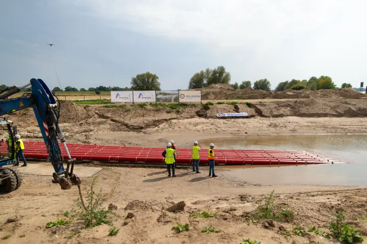 Ein Kran steht auf der linken Seite des Bildes. Ein langer, roter Rohrstrang, der in einer horizontalen Reihe auf dem Wasser treibt, erstreckt sich von der Uferkante in die Mitte. Am Ufer stehen drei Bauarbeiter in neongelben Westen und Schutzhelmen. Der Himmel ist bewölkt, mit einigen blauen Flecken. Im Hintergrund sind aufgeschüttete Erdhügel und Vegetation zu sehen. Ein Werbeschild mit den Logos von Amprion und Hülskens steht auf den Erdhügeln.