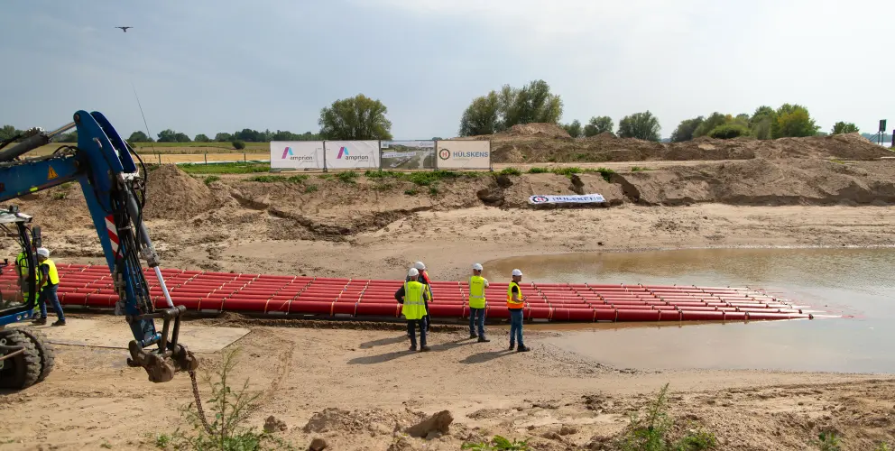 Ein Kran steht auf der linken Seite des Bildes. Ein langer, roter Rohrstrang, der in einer horizontalen Reihe auf dem Wasser treibt, erstreckt sich von der Uferkante in die Mitte. Am Ufer stehen drei Bauarbeiter in neongelben Westen und Schutzhelmen. Der Himmel ist bewölkt, mit einigen blauen Flecken. Im Hintergrund sind aufgeschüttete Erdhügel und Vegetation zu sehen. Ein Werbeschild mit den Logos von Amprion und Hülskens steht auf den Erdhügeln.
