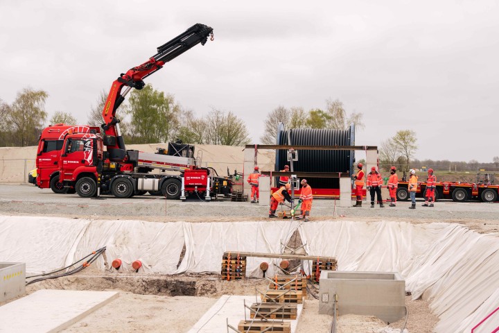 Das Bild zeigt eine Baustelle unter einem grauen, bewölkten Himmel. Im Vordergrund ist eine ausgehobene Grube zu sehen, deren Wände mit einer weißen Plane ausgekleidet sind. In der Grube befinden sich mehrere vorgefertigte Betonteile und Holzpaletten. Arbeiter in orangefarbenen Sicherheitswesten und Helmen sind in und um die Grube tätig.