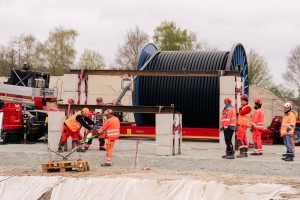 Das Bild zeigt eine Baustellenszene mit mehreren Bauarbeitern in orangefarbenen Warnwesten und Schutzhelmen. Sie sind mit verschiedenen Aufgaben beschäftigt.
Im Zentrum des Bildes befindet sich eine große, schwarze Kabeltrommel, die auf einer Achse gelagert ist und von einem weißen Gerüst gehalten wird. Links davon sind zwei Arbeiter zu sehen, die sich über etwas beugen, das auf einer Holzpalette liegt. Einer der Arbeiter trägt zusätzlich eine schwarze Jacke über seiner Warnweste.
Rechts von der Kabeltrommel stehen fünf weitere Arbeiter. Sie scheinen die Arbeiten zu beobachten oder Anweisungen zu geben. Einer dieser Arbeiter trägt einen weißen Schutzhelm, während die anderen rote Helme tragen.
Im Hintergrund sind Baumaschinen und -fahrzeuge teilweise verdeckt zu erkennen, darunter ein roter Kranwagen auf der linken Seite. Der Boden ist mit hellem Schotter bedeckt. Im hinteren Bereich sind Bäume ohne Blätter und ein trüber, grauer Himmel zu sehen, was auf eine kühle oder frühe Jahreszeit hindeutet. 