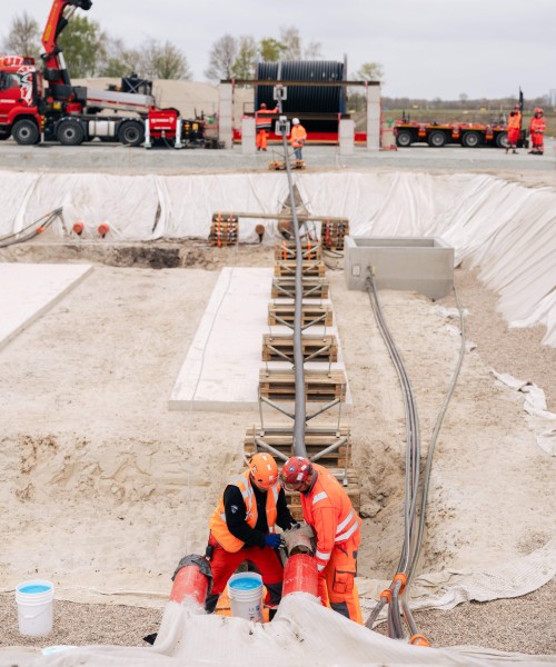 Das Bild zeigt eine weitläufige Baustelle unter einem bedeckten, hellgrauen Himmel. Im Vordergrund sind drei Bauarbeiter zu sehen, die sich über eine Reihe von orangefarbenen Rohren beugen, die in einem Graben verlegt sind. Die Arbeiter tragen orangefarbene Warnkleidung und Schutzhelme in Orange und Weiß. Einer der Arbeiter hat zusätzlich eine schwarze Weste über seiner Warnkleidung. Sie scheinen gerade Verbindungen an den Rohren zu prüfen oder zu bearbeiten.