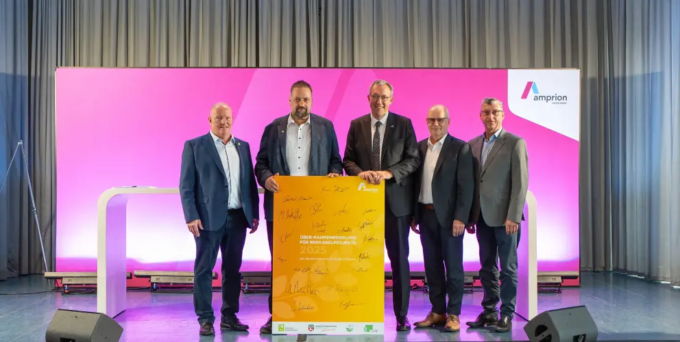 Fünf Männer in Anzügen stehen auf einer Bühne und halten gemeinsam ein gelbes Plakat mit Unterschriften, das die neue Rahmenregelung für Erdkabelprojekte 2025 zeigt. Der Hintergrund ist magentafarben mit Amprion-Logo.