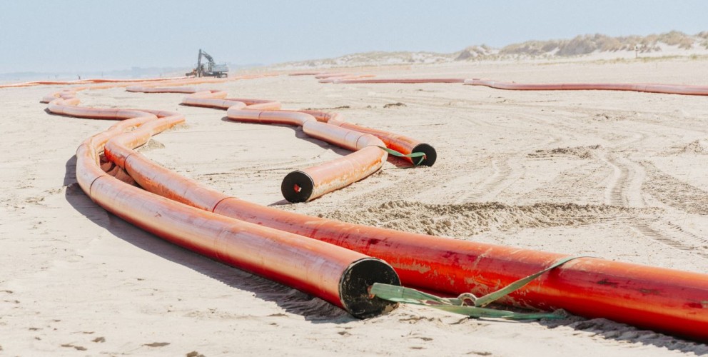 Das Bild zeigt eine weite, flache Sandlandschaft unter einem hellen Himmel, wahrscheinlich ein Strand oder eine Dünenlandschaft. Im Vordergrund und in den Mittelgrund erstrecken sich mehrere leuchtend orangefarbene Rohre, die sich schlängelnd über den Sandboden winden. Diese Rohre sind dick und scheinen aus Kunststoff zu sein, möglicherweise Kabelschutzrohre. Einige der Rohre sind miteinander verbunden, andere liegen lose nebeneinander oder parallel zueinander. An den Enden einiger Rohre sind schwarze, runde Öffnungen zu erkennen. Grüne Bänder oder Gurte sind an einigen Stellen um die Rohre gebunden, möglicherweise zur Sicherung oder zum Transport. Im Hintergrund, weiter entfernt auf dem Sand, ist schemenhaft ein großes Baufahrzeug, möglicherweise ein Bagger, zu sehen. Die gesamte Szene vermittelt den Eindruck einer Baustelle für Infrastrukturprojekte in einer Küstenregion.