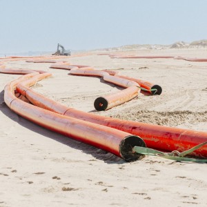 Das Bild zeigt eine weite, flache Sandlandschaft unter einem hellen Himmel, wahrscheinlich ein Strand oder eine Dünenlandschaft. Im Vordergrund und in den Mittelgrund erstrecken sich mehrere leuchtend orangefarbene Rohre, die sich schlängelnd über den Sandboden winden. Diese Rohre sind dick und scheinen aus Kunststoff zu sein, möglicherweise Kabelschutzrohre. Einige der Rohre sind miteinander verbunden, andere liegen lose nebeneinander oder parallel zueinander. An den Enden einiger Rohre sind schwarze, runde Öffnungen zu erkennen. Grüne Bänder oder Gurte sind an einigen Stellen um die Rohre gebunden, möglicherweise zur Sicherung oder zum Transport. Im Hintergrund, weiter entfernt auf dem Sand, ist schemenhaft ein großes Baufahrzeug, möglicherweise ein Bagger, zu sehen. Die gesamte Szene vermittelt den Eindruck einer Baustelle für Infrastrukturprojekte in einer Küstenregion.
