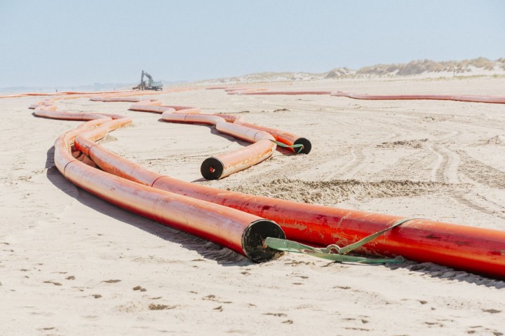 Das Bild zeigt eine weite, flache Sandlandschaft unter einem hellen Himmel, wahrscheinlich ein Strand oder eine Dünenlandschaft. Im Vordergrund und in den Mittelgrund erstrecken sich mehrere leuchtend orangefarbene Rohre, die sich schlängelnd über den Sandboden winden. Diese Rohre sind dick und scheinen aus Kunststoff zu sein, möglicherweise Kabelschutzrohre. Einige der Rohre sind miteinander verbunden, andere liegen lose nebeneinander oder parallel zueinander. An den Enden einiger Rohre sind schwarze, runde Öffnungen zu erkennen. Grüne Bänder oder Gurte sind an einigen Stellen um die Rohre gebunden, möglicherweise zur Sicherung oder zum Transport. Im Hintergrund, weiter entfernt auf dem Sand, ist schemenhaft ein großes Baufahrzeug, möglicherweise ein Bagger, zu sehen. Die gesamte Szene vermittelt den Eindruck einer Baustelle für Infrastrukturprojekte in einer Küstenregion.