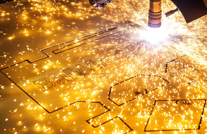 Das Bild zeigt eine Nahaufnahme eines Laserschneiders oder Plasmaschneiders, der in eine goldfarbene Metallplatte schneidet. Ein heller, funkelnder Lichtbogen und zahlreiche leuchtende Funken sprühen von der Schnittstelle. Das intensive Licht dominiert die rechte Seite des Bildes. Die Metallplatte ist horizontal ausgerichtet und weist bereits teilweise ausgeschnittene Formen auf, darunter der Text "BalWin". Der Hintergrund ist unscharf und dunkel, wodurch der Fokus vollständig auf den Schneidevorgang und die Funken gelegt wird. Die Szene vermittelt den Eindruck eines industriellen Fertigungsprozesses mit hoher Präzision und Energie.
English:
The image is a close-up shot of a laser or plasma cutter in action, cutting into a gold-colored metal sheet. A bright, sparkling arc and numerous glowing sparks emanate from the cutting interface. The intense light dominates the right side of the image. The metal plate is oriented horizontally and already shows partially cut-out shapes, including the text "BalWin 3". The background is blurred and dark, putting the entire focus on the cutting process and the sparks. The scene conveys the impression of an industrial manufacturing process characterized by high precision and energy.