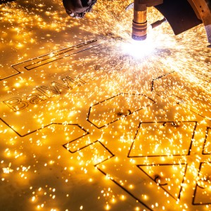 Das Bild zeigt eine Nahaufnahme eines Laserschneiders oder Plasmaschneiders, der in eine goldfarbene Metallplatte schneidet. Ein heller, funkelnder Lichtbogen und zahlreiche leuchtende Funken sprühen von der Schnittstelle. Das intensive Licht dominiert die rechte Seite des Bildes. Die Metallplatte ist horizontal ausgerichtet und weist bereits teilweise ausgeschnittene Formen auf, darunter der Text "BalWin". Der Hintergrund ist unscharf und dunkel, wodurch der Fokus vollständig auf den Schneidevorgang und die Funken gelegt wird. Die Szene vermittelt den Eindruck eines industriellen Fertigungsprozesses mit hoher Präzision und Energie.
English:
The image is a close-up shot of a laser or plasma cutter in action, cutting into a gold-colored metal sheet. A bright, sparkling arc and numerous glowing sparks emanate from the cutting interface. The intense light dominates the right side of the image. The metal plate is oriented horizontally and already shows partially cut-out shapes, including the text "BalWin". The background is blurred and dark, putting the entire focus on the cutting process and the sparks. The scene conveys the impression of an industrial manufacturing process characterized by high precision and energy.