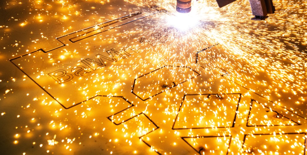Das Bild zeigt eine Nahaufnahme eines Laserschneiders oder Plasmaschneiders, der in eine goldfarbene Metallplatte schneidet. Ein heller, funkelnder Lichtbogen und zahlreiche leuchtende Funken sprühen von der Schnittstelle. Das intensive Licht dominiert die rechte Seite des Bildes. Die Metallplatte ist horizontal ausgerichtet und weist bereits teilweise ausgeschnittene Formen auf, darunter der Text "BalWin". Der Hintergrund ist unscharf und dunkel, wodurch der Fokus vollständig auf den Schneidevorgang und die Funken gelegt wird. Die Szene vermittelt den Eindruck eines industriellen Fertigungsprozesses mit hoher Präzision und Energie.
English:
The image is a close-up shot of a laser or plasma cutter in action, cutting into a gold-colored metal sheet. A bright, sparkling arc and numerous glowing sparks emanate from the cutting interface. The intense light dominates the right side of the image. The metal plate is oriented horizontally and already shows partially cut-out shapes, including the text "BalWin". The background is blurred and dark, putting the entire focus on the cutting process and the sparks. The scene conveys the impression of an industrial manufacturing process characterized by high precision and energy.