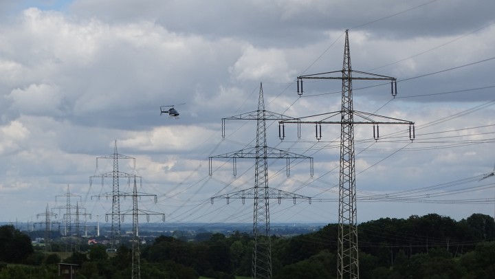 Das Bild zeigt eine Landschaft unter einem bewölkten Himmel, dominiert von mehreren großen Hochspannungsmasten, die sich in einer Reihe in die Ferne erstrecken. Diese Gittermasten aus Metall tragen zahlreiche Stromleitungen, die sich quer durch das Bild ziehen. Im linken Bildbereich, in mittlerer Entfernung, ist ein kleiner, dunkler Hubschrauber am Himmel zu sehen, der offenbar eine Freileitungskontrolle durchführt oder sich in deren Nähe befindet. Unterhalb der Masten und Leitungen erstreckt sich ein Waldgebiet mit Baumkronen, das im Hintergrund in eine Bebauung übergeht, die aber nur schemenhaft erkennbar ist.