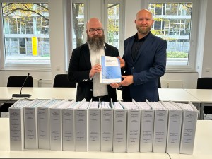 Zwei Männer in Anzügen stehen hinter einer langen Reihe von etwa 20 weißen Aktenordnern auf einem Konferenztisch und halten das Deckblatt eines Dokumentenstapels mit der Aufschrift 'Planfeststellungsunterlagen