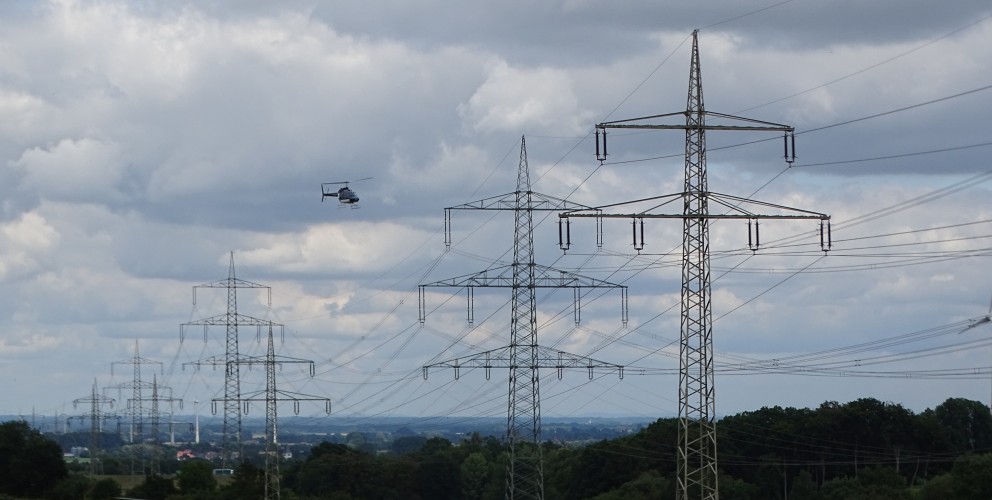 Beschreibung: Eine Aufnahme bei Tageslicht zeigt im Vordergrund zwei große Stahlgittermasten für Hochspannungsfreileitungen. Mehrere Stromkabel spannen sich in den grauen, bewölkten Himmel. Links im Bild fliegt ein kleiner, dunkler Helikopter. Im mittleren und hinteren Bereich erstreckt sich eine grüne, bewaldete Hügellandschaft, dahinter sind diffuse Umrisse einer Industriezone oder Stadt zu erkennen.