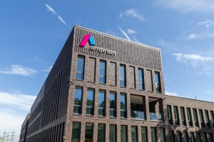 Moderne Außenansicht eines mehrstöckigen Bürogebäudes mit Klinkerfassade und großem Amprion-Logo an der Ecke des Gebäudes. Das Logo besteht aus einem pink-blauen grafischen Element und dem Schriftzug „amprion“ in Kleinbuchstaben. Der Himmel ist klar und blau, einige weiße Wolken ziehen vorbei. Das Gebäude wirkt neu und repräsentativ.