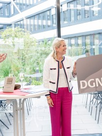 Drei Personen in einem Innenbereich mit großen Glasfenstern. Auf einem Stehtisch steht eine Platin-Auszeichnungsplakette der DGNB. Der Mann links applaudiert, die Frau in der Mitte lächelt, der Mann rechts hält das große DGNB-Logo. Auf dem Tisch stehen auch Sektgläser, was auf eine feierliche Übergabe oder einen Empfang hinweist.