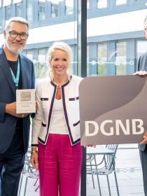 Drei Personen stehen nebeneinander in einem modernen, lichtdurchfluteten Gebäude mit Glasfassade. Der Mann links hält eine Plakette mit der Aufschrift „DGNB Platin“, die Frau in der Mitte lächelt, und der Mann rechts hält ein großes Schild mit dem DGNB-Logo. Alle drei sind festlich gekleidet, die Szene wirkt feierlich. Es handelt sich offensichtlich um eine Auszeichnung im Bereich nachhaltiges Bauen oder Architektur.