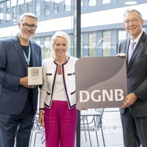 Drei Personen stehen nebeneinander in einem modernen, lichtdurchfluteten Gebäude mit Glasfassade. Der Mann links hält eine Plakette mit der Aufschrift „DGNB Platin“, die Frau in der Mitte lächelt, und der Mann rechts hält ein großes Schild mit dem DGNB-Logo. Alle drei sind festlich gekleidet, die Szene wirkt feierlich. Es handelt sich offensichtlich um eine Auszeichnung im Bereich nachhaltiges Bauen oder Architektur.