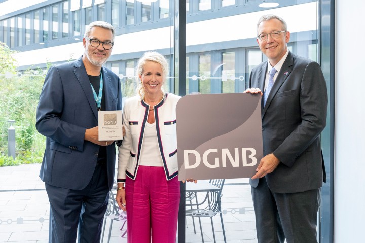 Drei Personen stehen nebeneinander in einem modernen, lichtdurchfluteten Gebäude mit Glasfassade. Der Mann links hält eine Plakette mit der Aufschrift „DGNB Platin“, die Frau in der Mitte lächelt, und der Mann rechts hält ein großes Schild mit dem DGNB-Logo. Alle drei sind festlich gekleidet, die Szene wirkt feierlich. Es handelt sich offensichtlich um eine Auszeichnung im Bereich nachhaltiges Bauen oder Architektur.
