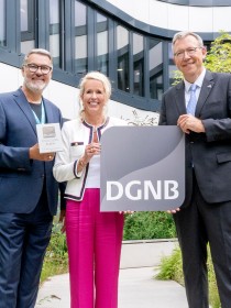 Drei Personen stehen im begrünten Innenhof eines modernen Bürogebäudes. Der Mann links hält eine Plakette mit der Aufschrift „DGNB Platin“, die Frau in der Mitte lächelt und berührt mit einer Hand das Schild mit dem DGNB-Logo, das der Mann rechts in einem dunklen Anzug präsentiert. Die Szene wirkt feierlich und symbolisiert eine Auszeichnung im Bereich nachhaltiges Bauen.