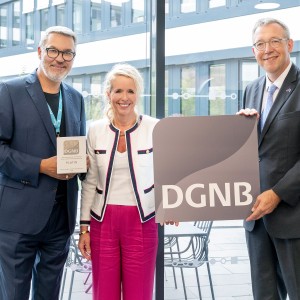Drei Personen stehen nebeneinander in einem modernen, lichtdurchfluteten Gebäude mit Glasfassade. Der Mann links hält eine Plakette mit der Aufschrift „DGNB Platin“, die Frau in der Mitte lächelt, und der Mann rechts hält ein großes Schild mit dem DGNB-Logo. Alle drei sind festlich gekleidet, die Szene wirkt feierlich. Es handelt sich offensichtlich um eine Auszeichnung im Bereich nachhaltiges Bauen oder Architektur.