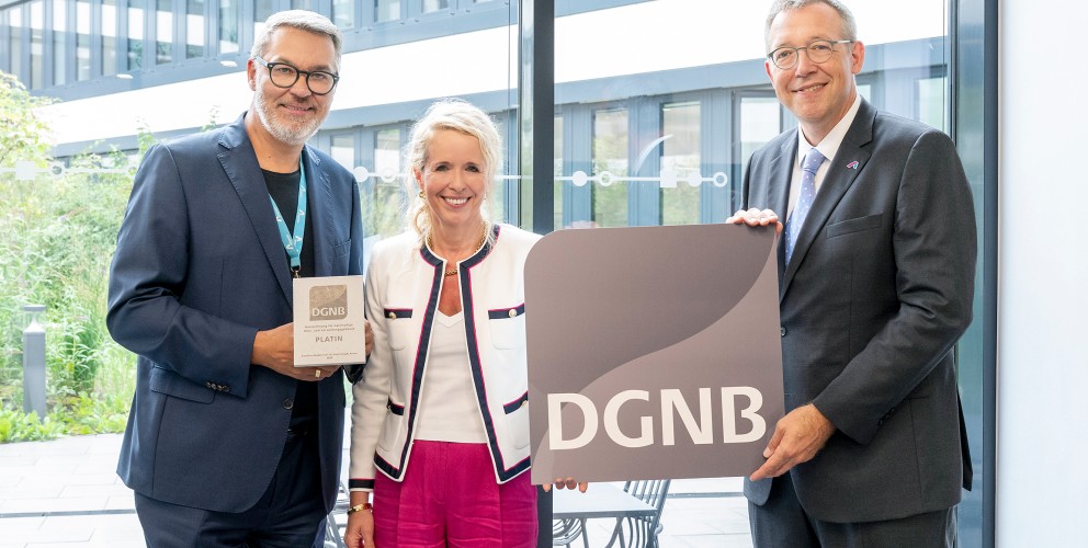 Drei Personen stehen nebeneinander in einem modernen, lichtdurchfluteten Gebäude mit Glasfassade. Der Mann links hält eine Plakette mit der Aufschrift „DGNB Platin“, die Frau in der Mitte lächelt, und der Mann rechts hält ein großes Schild mit dem DGNB-Logo. Alle drei sind festlich gekleidet, die Szene wirkt feierlich. Es handelt sich offensichtlich um eine Auszeichnung im Bereich nachhaltiges Bauen oder Architektur.