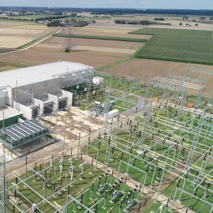 Das Bild zeigt eine Konverterstation aus der Vogelperspektive. Im Zentrum befindet sich ein großes, rechteckiges Gebäude mit einem hellgrauen Dach und hellgrauen oberen Wandteilen. Die unteren Wandteile sind in einem dunklen Grün gehalten. An einer Seite des Hauptgebäudes schließt ein kleineres, ebenfalls rechteckiges Gebäude mit einem flachen, begrünten Dach an.
Um das Hauptgebäude herum befinden sich verschiedene technische Anlagen und Konstruktionen aus Stahlträgern. Besonders auffällig ist eine größere, quaderförmige Anlage mit mehreren Ebenen und zahlreichen grauen Komponenten. Leitungen und Kabel verlaufen zwischen den verschiedenen Anlagenteilen. Der Boden um die Gebäude und Anlagen ist größtenteils mit hellem Schotter bedeckt.
Im Vordergrund und auf der rechten Seite des Bildes erstreckt sich ein weitläufiges Freiluftschaltanlagengelände. Zahlreiche hohe, filigrane Stahlgittermasten tragen Isolatoren und elektrische Leiter. Die Anordnung wirkt komplex und strukturiert. Der Boden unter den Masten ist mit kurzem, grünem Gras bewachsen.
Im Hintergrund erstreckt sich eine ländliche Landschaft mit Feldern in verschiedenen Brauntönen und Grüntönen, die wie ein Schachbrettmuster angeordnet sind. Vereinzelt sind Bäume und Sträucher zu erkennen. Am Horizont ist eine Baumreihe und darüber ein heller, leicht bewölkter Himmel sichtbar. Eine einzelne Hochspannungsleitung mit einem markanten Gittermast ist ebenfalls im Hintergrund zu sehen und führt über die Felder.
