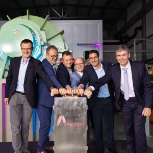 Sechs Männer in Anzügen stehen nebeneinander in einer Industriehalle vor einer großen technischen Anlage mit grünem Zylindergehäuse. Gemeinsam drücken sie symbolisch auf einen metallischen Startknopf mit dem Amprion-Logo. Der Raum ist technisch ausgestattet, mit sichtbaren Leitungen und Maschinen im Hintergrund. Die Szene symbolisiert die feierliche Inbetriebnahme einer Anlage, vermutlich im Kontext der Energiewende.