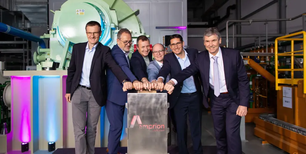 Sechs Männer in Anzügen stehen nebeneinander in einer Industriehalle vor einer großen technischen Anlage mit grünem Zylindergehäuse. Gemeinsam drücken sie symbolisch auf einen metallischen Startknopf mit dem Amprion-Logo. Der Raum ist technisch ausgestattet, mit sichtbaren Leitungen und Maschinen im Hintergrund. Die Szene symbolisiert die feierliche Inbetriebnahme einer Anlage, vermutlich im Kontext der Energiewende.