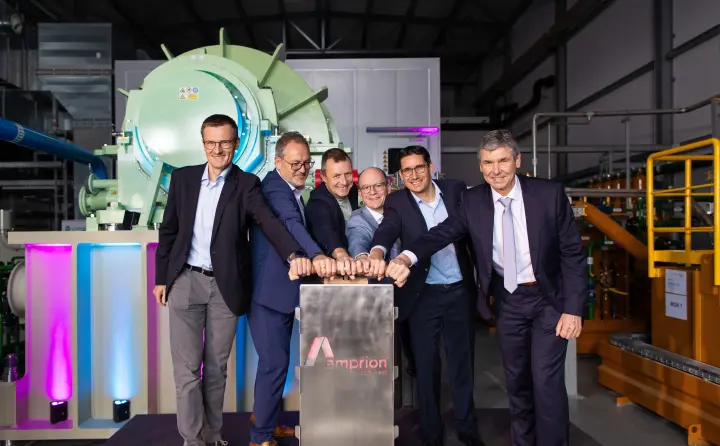Sechs Männer in Anzügen stehen nebeneinander in einer Industriehalle vor einer großen technischen Anlage mit grünem Zylindergehäuse. Gemeinsam drücken sie symbolisch auf einen metallischen Startknopf mit dem Amprion-Logo. Der Raum ist technisch ausgestattet, mit sichtbaren Leitungen und Maschinen im Hintergrund. Die Szene symbolisiert die feierliche Inbetriebnahme einer Anlage, vermutlich im Kontext der Energiewende.