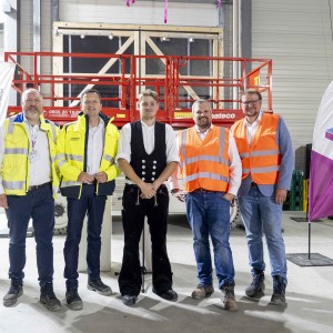 Fünf Männer stehen in einer Halle vor einer roten Scherenbühne. Zwei von ihnen tragen gelbe Sicherheitswesten, zwei orangefarbene und einer ist in traditioneller schwarzer Arbeitskleidung gekleidet. Im Hintergrund sind Werbefahnen und die Wände der Halle zu sehen.
