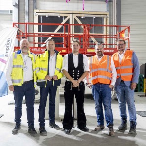 Fünf Männer stehen in einer Halle vor einer roten Scherenbühne. Zwei von ihnen tragen gelbe Sicherheitswesten, zwei orangefarbene und einer ist in traditioneller schwarzer Arbeitskleidung gekleidet. Im Hintergrund sind Werbefahnen und die Wände der Halle zu sehen.