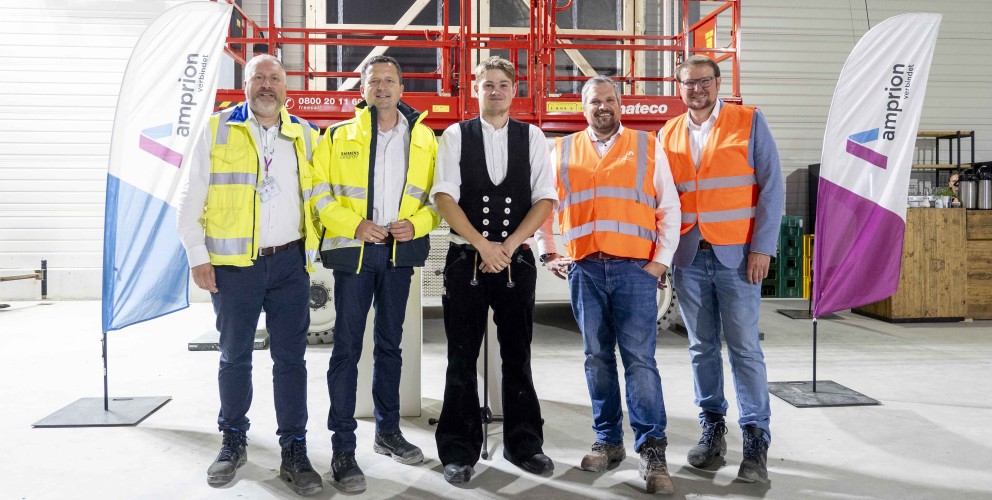 Fünf Männer stehen in einer Halle vor einer roten Scherenbühne. Zwei von ihnen tragen gelbe Sicherheitswesten, zwei orangefarbene und einer ist in traditioneller schwarzer Arbeitskleidung gekleidet. Im Hintergrund sind Werbefahnen und die Wände der Halle zu sehen.
