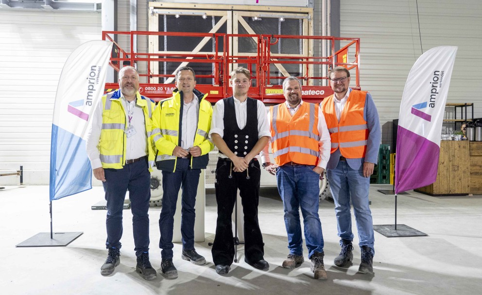Fünf Männer stehen in einer Halle vor einer roten Scherenbühne. Zwei von ihnen tragen gelbe Sicherheitswesten, zwei orangefarbene und einer ist in traditioneller schwarzer Arbeitskleidung gekleidet. Im Hintergrund sind Werbefahnen und die Wände der Halle zu sehen.