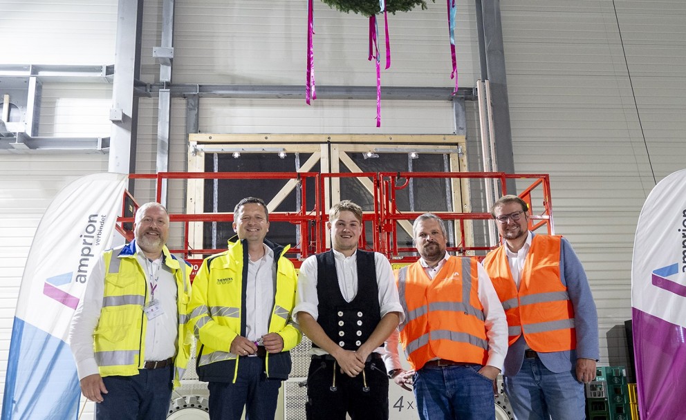 Auf dem Foto stehen fünf Männer in einer Industriehalle. In der Mitte der Gruppe hängt von der Decke ein geschmückter Kranz. Die Männer blicken lächelnd in die Kamera.
