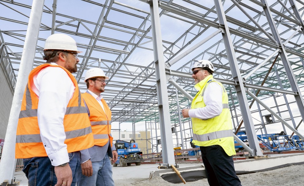 Vordergrund
Drei Männer, die auf einer Baustelle stehen und miteinander reden. Sie tragen Schutzhelme und Sicherheitswesten.
Links steht ein Mann im Profil, der eine orangefarbene Weste und Jeans trägt.
In der Mitte steht ein Mann, ebenfalls im Profil, mit orangefarbener Weste, Sakko und Jeans. Er trägt eine Brille und einen Schutzhelm.
Rechts steht ein Mann mit Brille und Bart in einer gelben Sicherheitsweste. Er hat seinen Blick der Mitte zugewandt und gestikuliert mit den Händen.
Hintergrund
Die Männer befinden sich in einer großen, noch nicht fertiggestellten Industriehalle. Die Stahlkonstruktion ist das dominierende Element im Hintergrund, mit zahlreichen silbernen Trägern, die das Dachgerüst bilden. Der Boden ist noch unbefestigt, teilweise mit Kies bedeckt. Im Hintergrund ist eine blaue Baumaschine zu sehen.