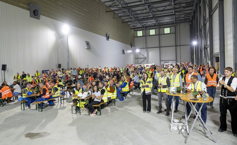 Das Foto zeigt eine große Gruppe von Menschen, die sich in einer riesigen Industriehalle versammelt haben. Viele von ihnen tragen gelbe oder orangefarbene Sicherheitswesten. Im Vordergrund klatschen einige Personen.
Details
Sitzplätze: Die meisten Anwesenden sitzen auf Bierbänken an einfachen Holztischen. Auf den Tischen stehen Getränke und Speisen.
Stehplätze: Rechts im Bild steht eine Gruppe von Männern um einen runden Stehtisch.
Halle: Die Wände sind hellgrau und die Deckenkonstruktion besteht aus großen Stahlträgern. Ein paar Lüftungsrohre und Strahler sind an den Wänden und der Decke zu sehen.
Kleidung: Viele Teilnehmer sind in Arbeitskleidung gekleidet, einige tragen weiße Schutzhelme. Einer der Männer im Vordergrund ist in traditioneller Zimmermannskleidung gekleidet, was auf ein Richtfest hindeutet.