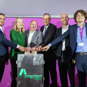 Fünf Männer und eine Frau in Geschäftskleidung legen einen Schalter um. Der Schalter befindet sich auf einem Aufsteller mit Amprion-Logo. Im Hintergrund eine fuchsiafarbene Pressewand.