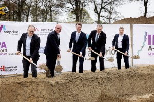 Das Foto zeigt fünf Männer in Anzügen, die an einem Spatenstich teilnehmen. Sie stehen nebeneinander an einer länglichen, aufgeworfenen Erdgrube und halten goldfarbene Spaten in den Händen, mit denen sie Erde bewegen. Im Hintergrund ist ein weißes Banner mit verschiedenen Logos und Schriftzügen zu sehen. Links ist das Amprion-Logo mit dem Schriftzug "amprion verbindet" erkennbar. Daneben steht "HEITKAMP" mit dem Untertitel "Baumannschaften. Unsere Welt. Unsere Leistung.". Rechts sind Teile eines weiteren Logos mit "A-" und "GEM" sichtbar. Hinter dem Banner befinden sich Bäume ohne Blätter und ein sandiger oder erdfarbener Hügel. 