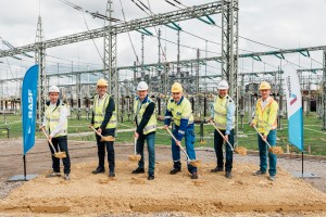 Sechs Männer in Schutzkleidung stehen vor einer Umspannanlage auf sandigem Untergrund. Neben ihnen befinden sich Fahnen von BASF und Amprion. Die Männer halten Spaten mit etwas Sand in den Händen und blicken direkt in die Kamera.