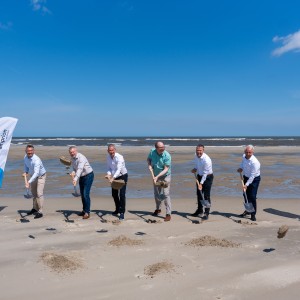 
Sechs Männer in Hemden und Hosen stehen nebeneinander auf einem Sandstrand und halten Spaten in den Händen, als würden sie etwas ausgraben. Sie blicken alle in Richtung des Betrachters. Hinter ihnen erstreckt sich das Meer unter einem klaren blauen Himmel mit wenigen Wolken. Links und rechts im Bild sind jeweils eine Fahne mit dem Logo "amprion" zu sehen. Die Fahne auf der linken Seite ist blau und weiß, die auf der rechten Seite ist violett und weiß.