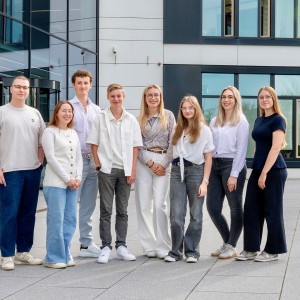  junge Menschen stehen für ein Foto im Freien. Sie stehen vor einem modernen Bürogebäude und tragen lockere Kleidung. 