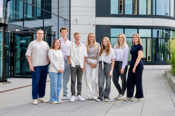  junge Menschen stehen für ein Foto im Freien. Sie stehen vor einem modernen Bürogebäude und tragen lockere Kleidung. 