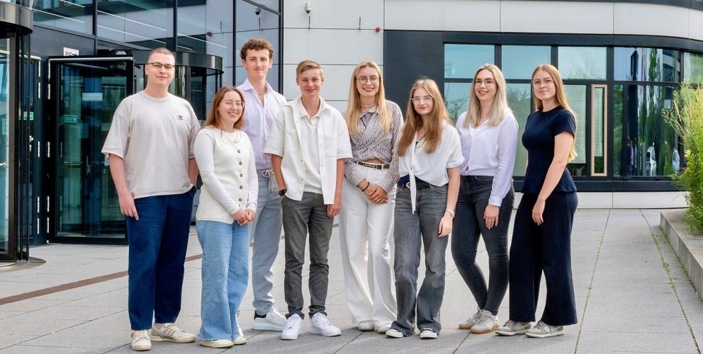  junge Menschen stehen für ein Foto im Freien. Sie stehen vor einem modernen Bürogebäude und tragen lockere Kleidung. 