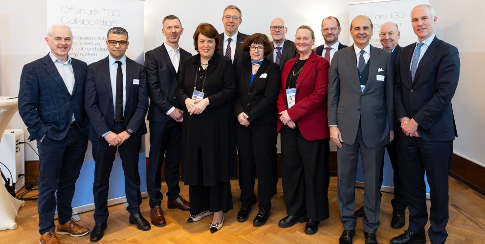 Gruppe von neun Personen in Businesskleidung vor weißen Stellwänden mit Text, stehend auf Parkettboden
