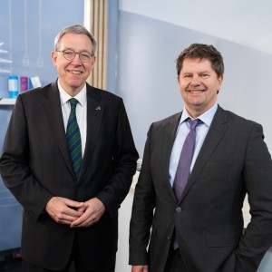 Zwei Männer in dunklen Anzügen stehen nebeneinander und lächeln in die Kamera. Im Hintergrund eine hellblaue Wand mit einer stilisierten Grafik eines Strommasten und einer Windkraftanlage. Bildnachweis: Amprion GmbH / Frank Peterschröder.