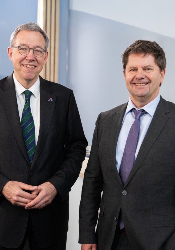 Zwei Männer in dunklen Anzügen stehen nebeneinander und lächeln in die Kamera. Im Hintergrund eine hellblaue Wand mit einer stilisierten Grafik eines Strommasten und einer Windkraftanlage. Bildnachweis: Amprion GmbH / Frank Peterschröder.