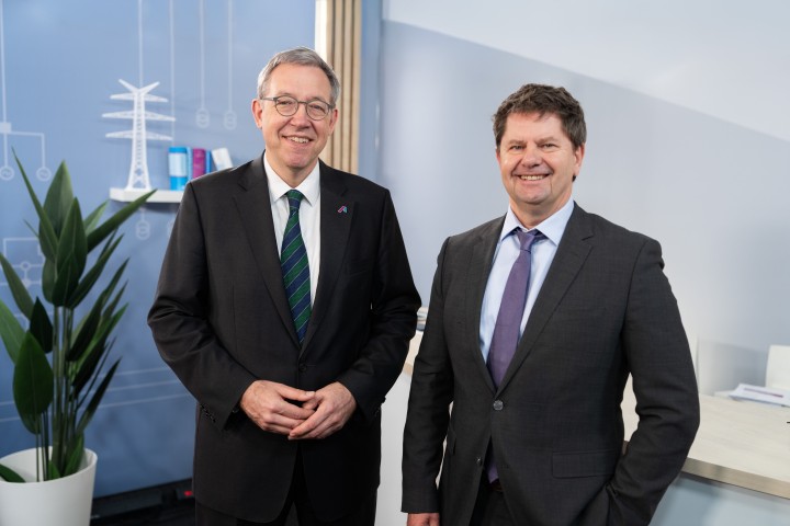 Zwei Männer in dunklen Anzügen stehen nebeneinander und lächeln in die Kamera. Im Hintergrund eine hellblaue Wand mit einer stilisierten Grafik eines Strommasten und einer Windkraftanlage. Bildnachweis: Amprion GmbH / Frank Peterschröder.
